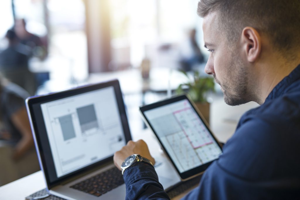 Mężczyzna w biurze software house analizuje projekt na laptopie i tablecie, koncentrując się na tworzeniu oprogramowania i planowaniu rozwoju aplikacji.